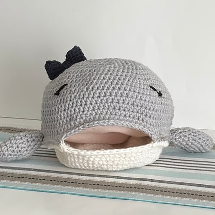 Large handmade crochet gray whale with its mouth open and a navy blue bow on its head.  You can see pink inside its open mouth and it is on a blue and gray striped surface with a white background