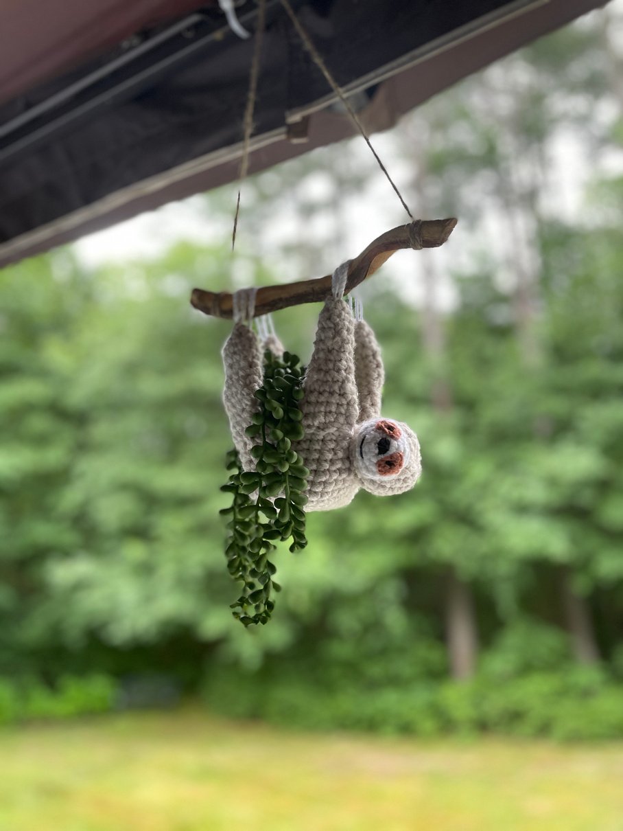 Handmade crochet sloth planter hanging on a branch by all four of its hands. The branch is hanging with twine and 
and the sloth has a string of pearls plant inside that hangs over the edge of its belly.  The background is blurred green trees.